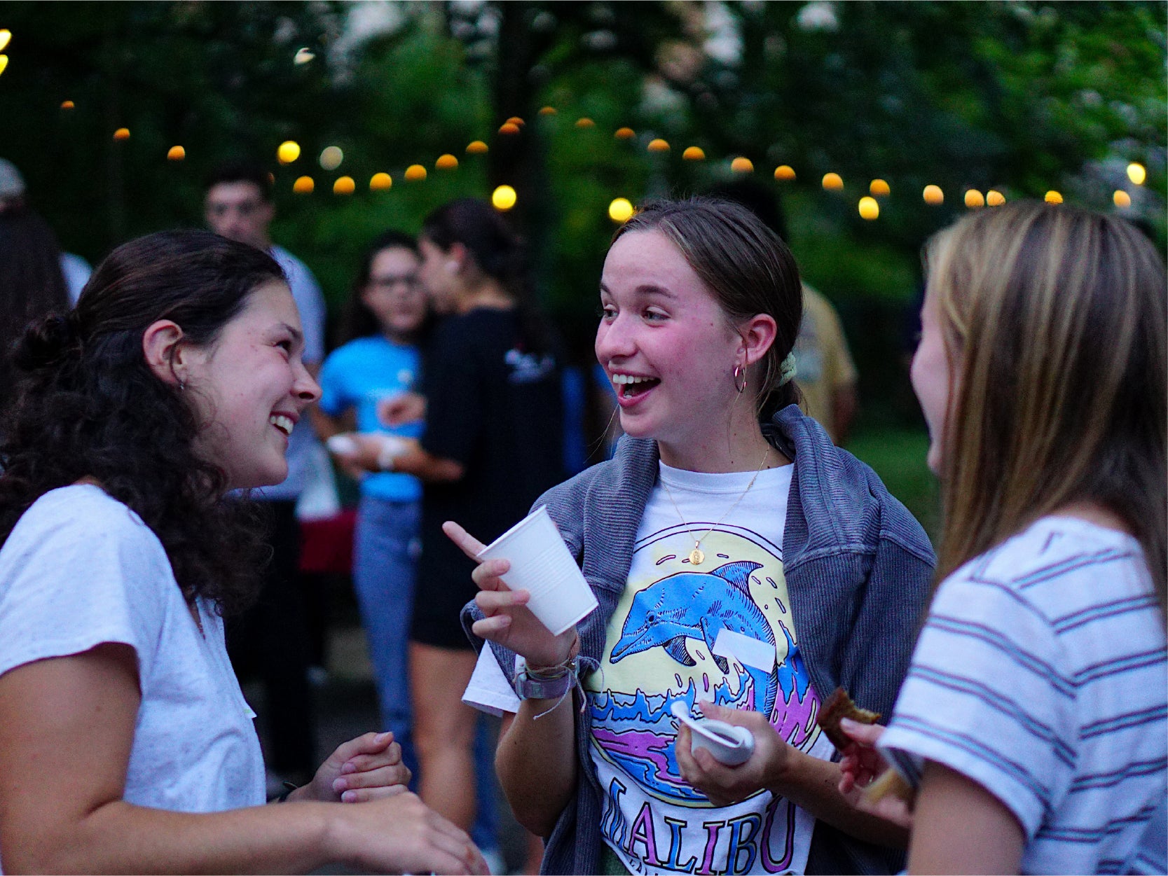 Three UMD students laugh and talk outside under ambient Edison string lights at dusk.