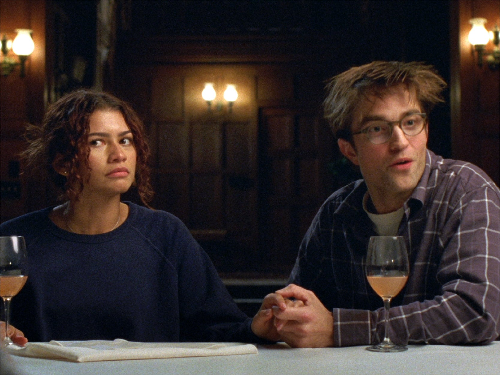 Zendaya and Robert Pattinson sitting at a table both with a wine glass and have a confused facial expression. This is a promotional image from The Drama.