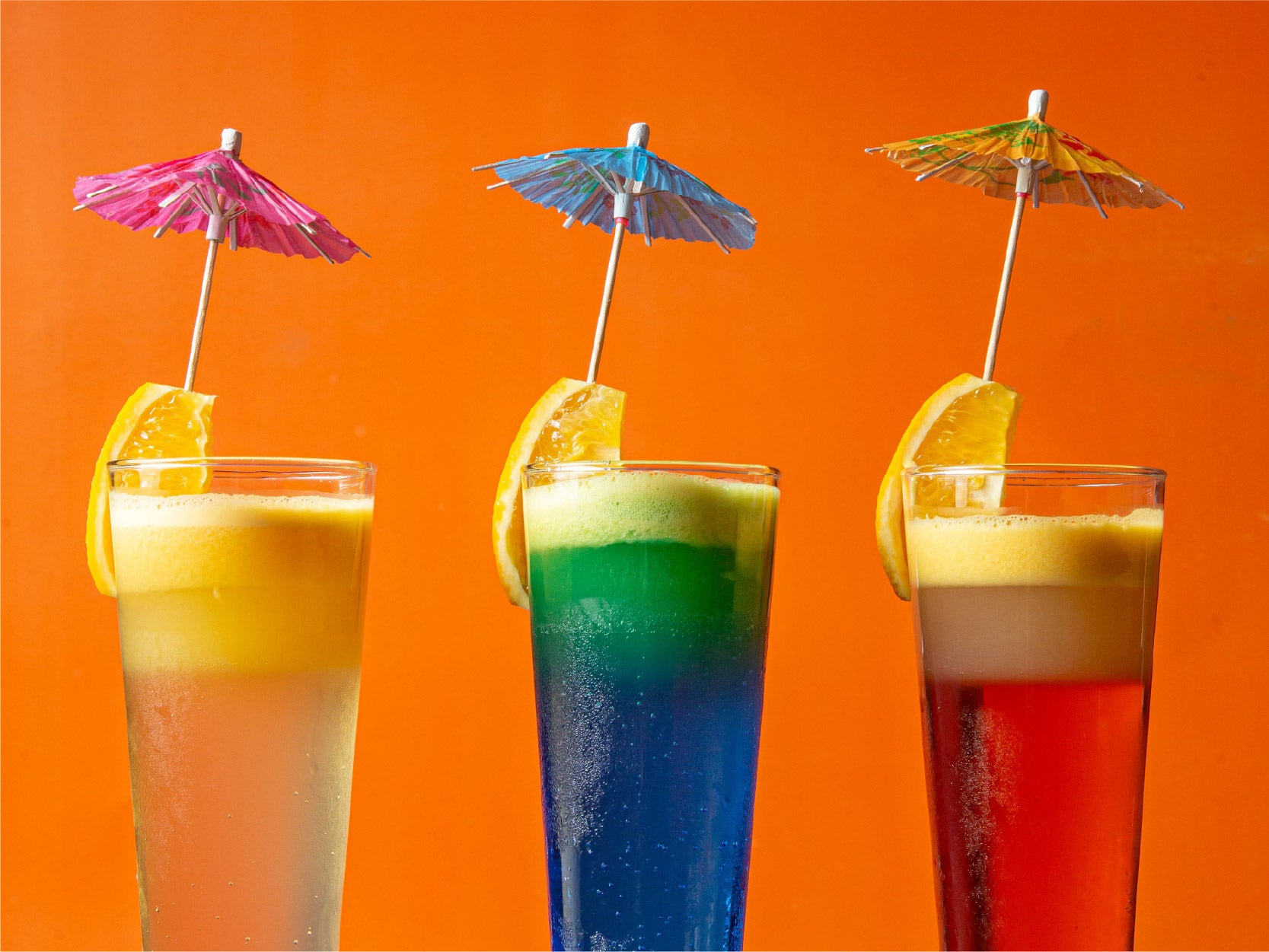 A image of 3 cups of colorful drinks all garnished with an orange and a mini umbrella.