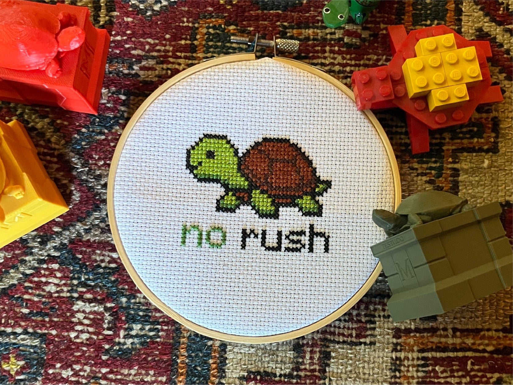 A photo of a turtle cross-stitch project with the text "no rush" at the bottom. The project is surrounded by turtle toys in various colors.